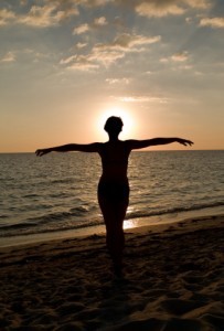 A woman standing on the beach, by the sea, watching a sunset.