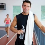 A man standing on an indoor running track with a bottle of water and a towel.