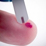 A finger with a spec of blood and a blood glucose monitoring strip.