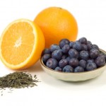 A bowl of blueberries next to a half orange, a whole orange and a pile of green tea.