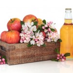 A glass bottle of cider next to a wooden crate of apples and flowers.