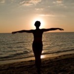 A woman standing on the beach, by the sea, watching a sunset.