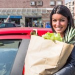 A woman standing next to a car with a bag of groceries.